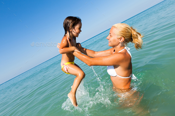 Young mother standing in blue water and playing with her smiling daughter