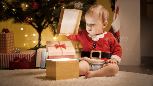 Cute baby boy sitting under Christmas tree and opening lid of golden gift box