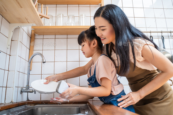 Asian loving mom teach young preschool girl to wash dishes in house.