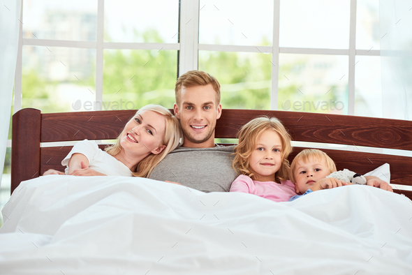 Family day. Young beautiful family of four people relaxing in bed together and looking at camera