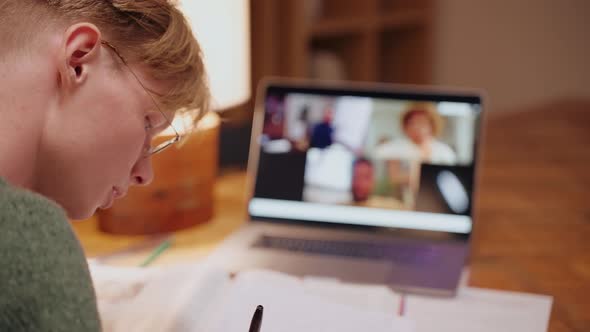 Young Man Studying at Home Having a Video Call on a Laptop Writing His Notes alt