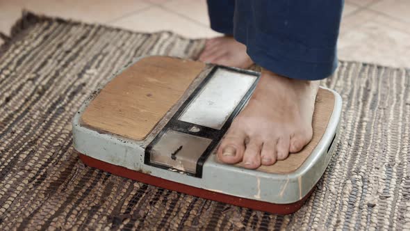 Senior sick woman stands on an old dirty scale and measures her weight. alt