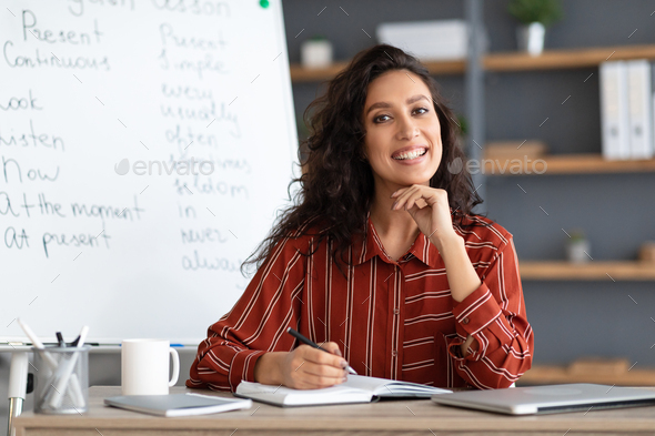 Female teacher sitting at desk looking at camera - Stock Photo - Images