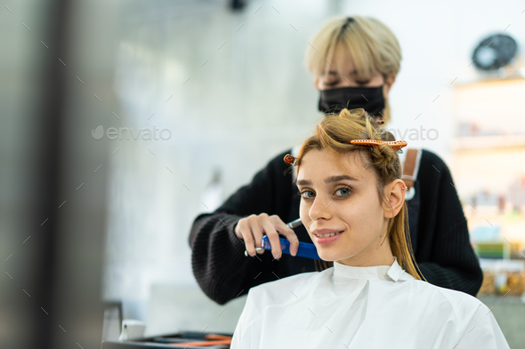 Asian female stylist wear mask and face shield cutting Caucasian woman\'s hair in salon.