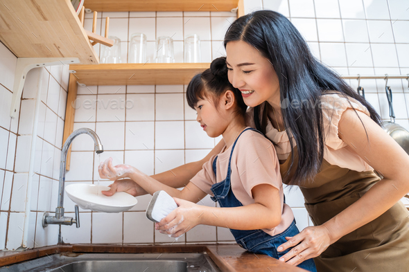 Asian loving mom teach young preschool girl to wash dishes in kitchen in house.