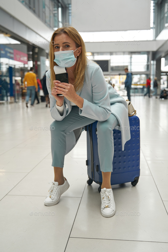 Low-spirited tourist in face mask sitting on her baggage