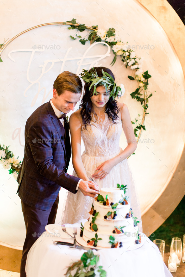The close-up portrait of the newlywed couple cutting the rustic wedding cake with figs and