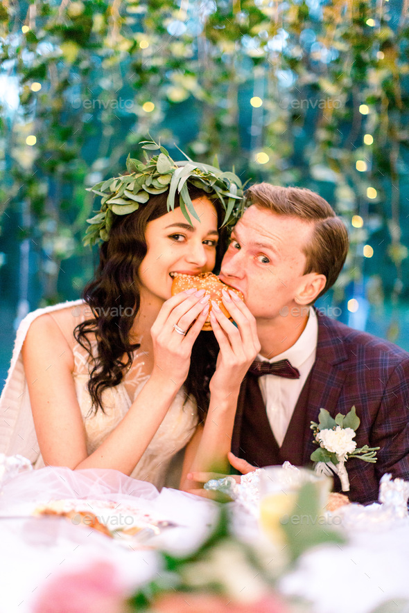 Romantic wedding couple eating a burger together at a rustic restaurant outdoors in forest, looking