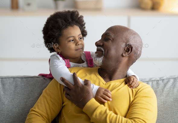 Cheerful little black girl play with older grandfather from back, hugging man at home interior