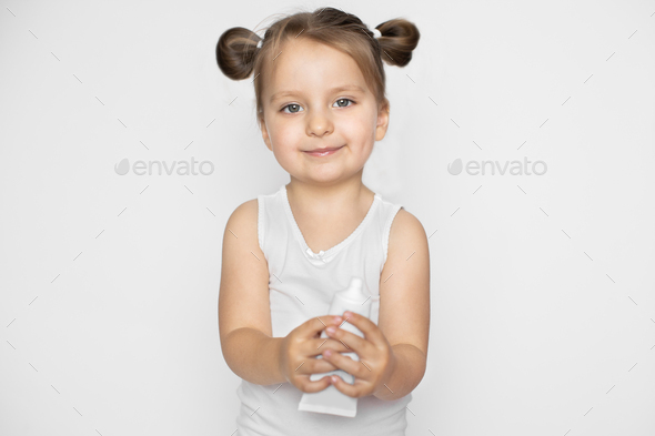 A little girl with blonde hair in two ponytails, wearing white top, holding tube of cream. Promo of
