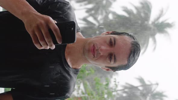 Vetrical Shot of Wet Young Man Standing in the Pouring Tropical Rain Using His Smartphone alt