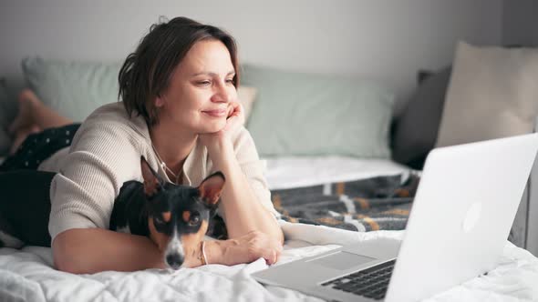 A Middleaged Woman Is Working on Her Laptop While Lying on the Bed with Her Dog alt