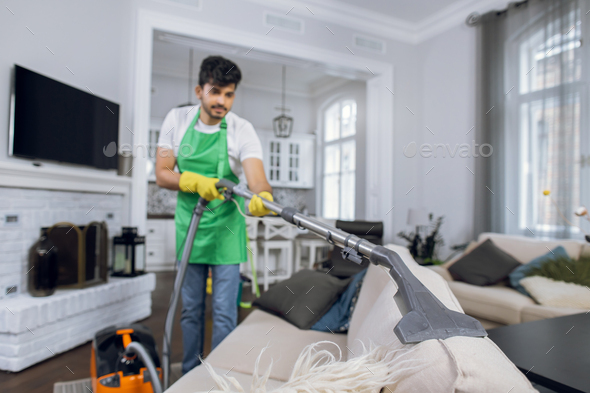 young indian man removing dust from sofa with vacuum cleaner