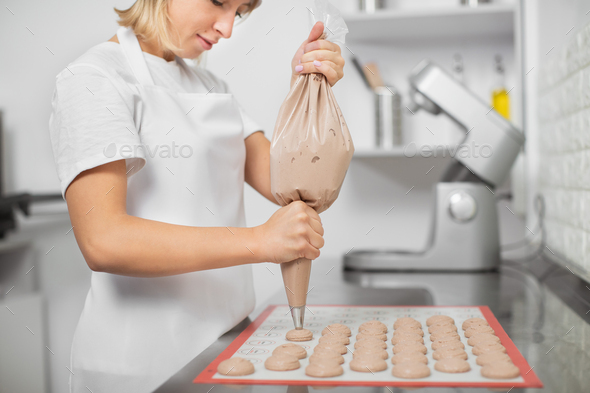 Female young confectioner making macarons in her cozy light pastry shop.