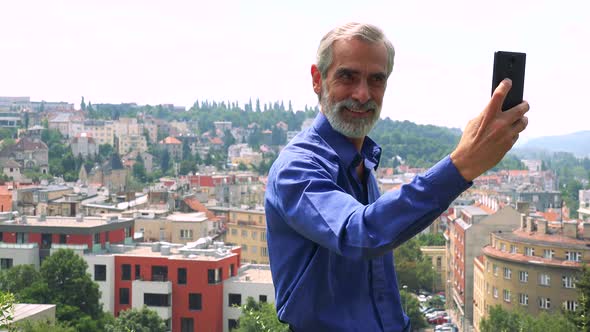 Old Senior Man Photographs with Smartphone (Selfie) - City (Buildings) in Background alt