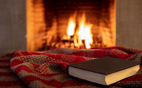 Cup of hot drink and book on blanket near fireplace indoors