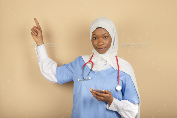 Muslim religion nurse online with headscarf .Holds smartphone, points finger up