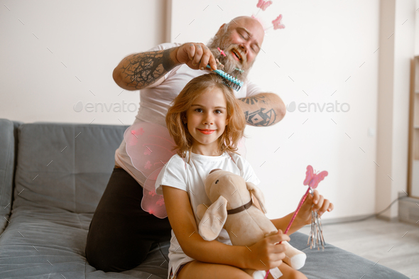 Smiling girl holds toy dog and magic stick while dad brushes her hair in room