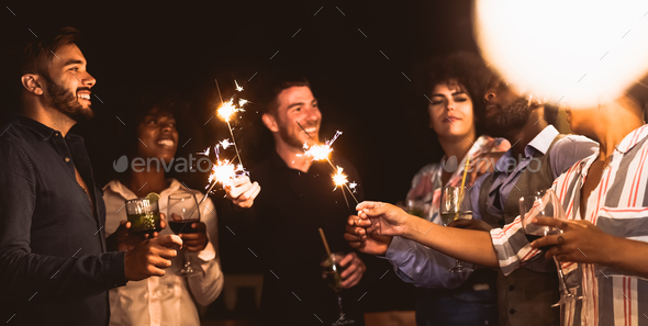 Happy young friends having fun with sparklers fireworks while drinking cocktails on house patio