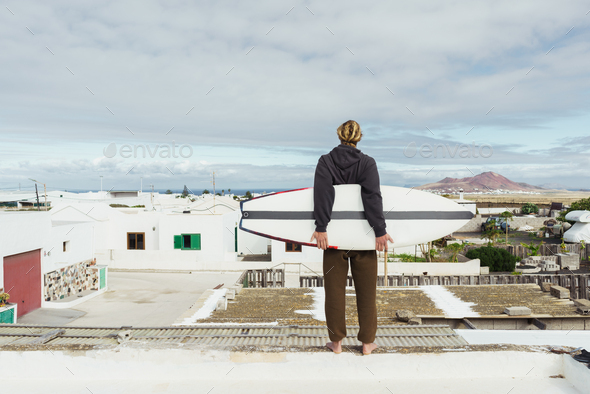 Man standing with surf board on roof of building and looking at mountain