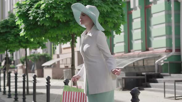 Portrait of Elegant Caucasian Woman Lost in Foreign City. Uncertain Stylish Lady with Shopping Bags alt