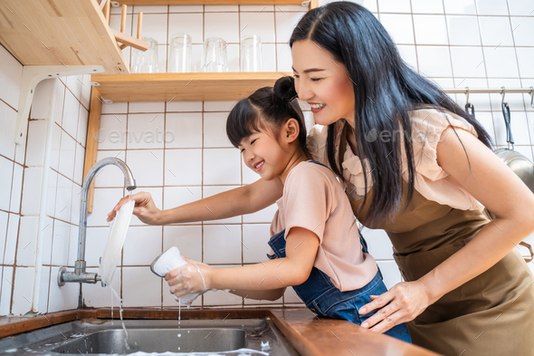 Asian mother teach little kid daughter wash dishes in kitchen in house.