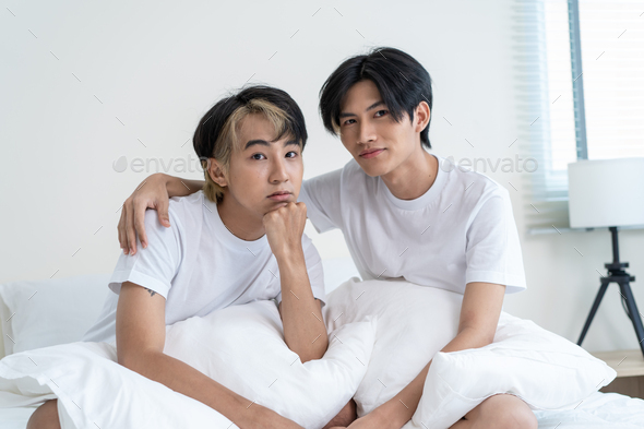 Portrait of Asian handsome gay couple sit on bed in bedroom and smile looking at camera in house.