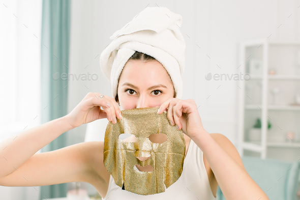 Young pretty woman in white towel after bath holding paper gold facial mask.