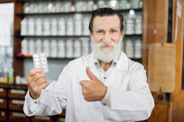 Smiling joyful senior male pharmacist in white coat, standing in ancient vintage pharmacy