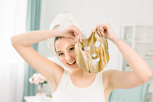 Young pretty woman in white towel after bath holding paper gold facial mask