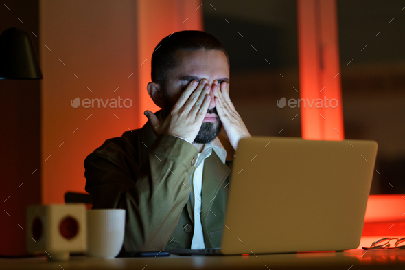 Exhausted web designer or manager work late overloaded alone in office use at laptop rubbing eyes