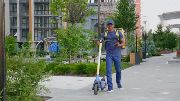 Afroamerican Man Courier Walks with Electric Scooter Uses Smartphone Navigate alt