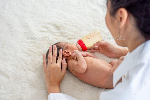 Asian mother give milk feeding with bottle to little newborn baby on white bed and take care