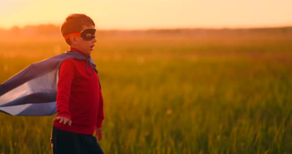 A Boy in a Superhero Costume at Sunset Runs Across the Field Laughing and Smiling alt