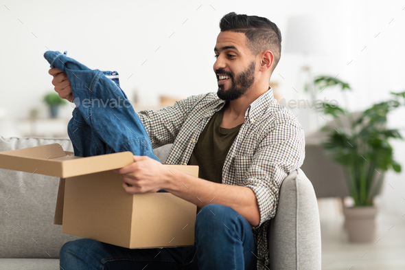Happy Arab man taking out new clothes from carton box, satisfied with delivery service at home
