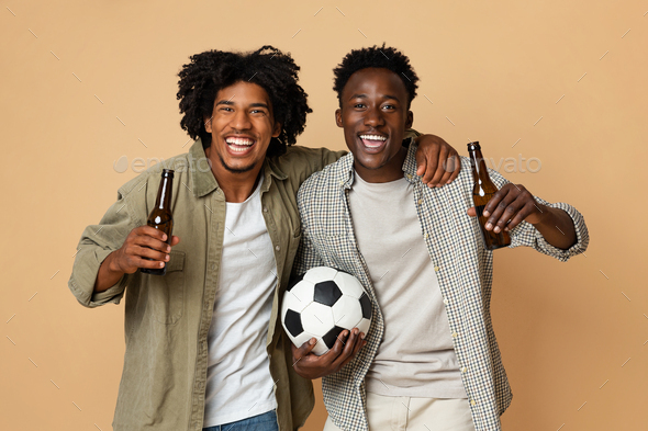 Football Fans. Two Cheerful Black Guys Holding Soccer Ball And Drinking Beer