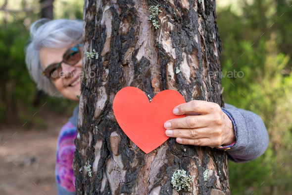 Senior woman in the woods hugs a tree trunk putting a heart shape on it. Earth day concept.