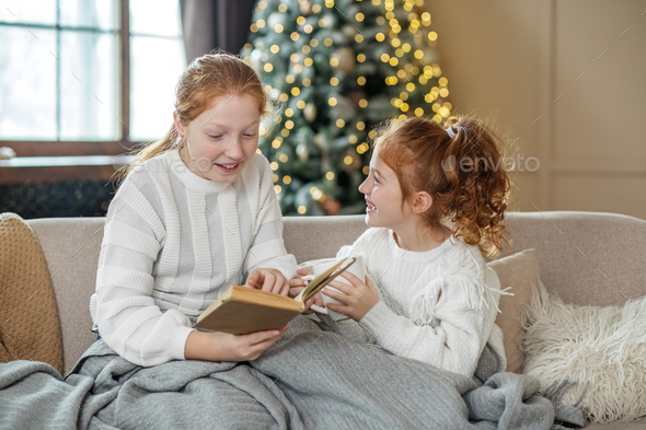 Two sisters are sitting on the couch and reading a book. Delicious hot cocoa. - Stock Photo - Images