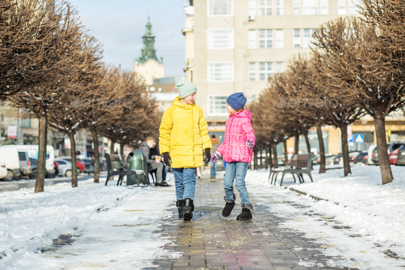 Two children talk and walk down the street in winter jackets and warm hats.