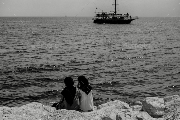 Back view of two women wearing the hijab sitting on rocky beach