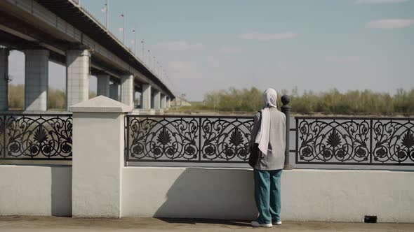 Muslin Woman with Long Hijab Stands By Fence on Waterfront alt