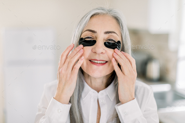 senior gray haired woman, applying under eye mask, black patches on her face
