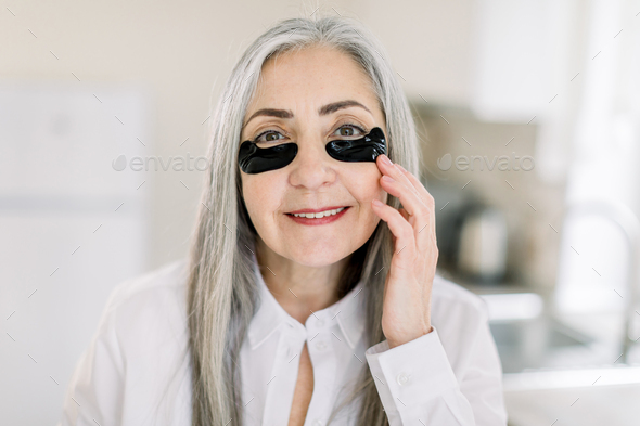 senior gray haired woman, applying under eye mask, black patches on her face with hands