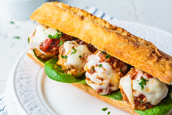 Sub meatballs sandwich with cheese and tomato sauce in baguette on white plate.