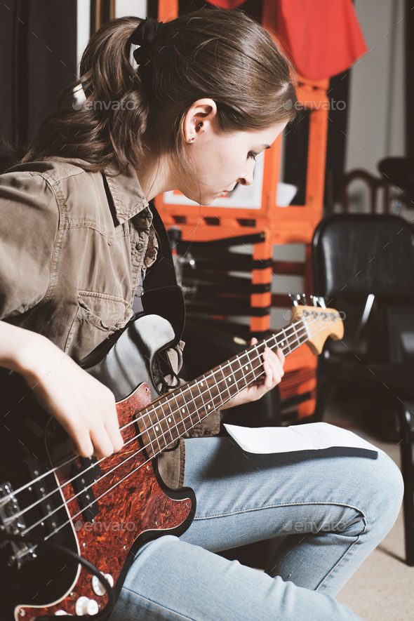 young woman playing bass guitar and looking at chords notes. rehearsal room.