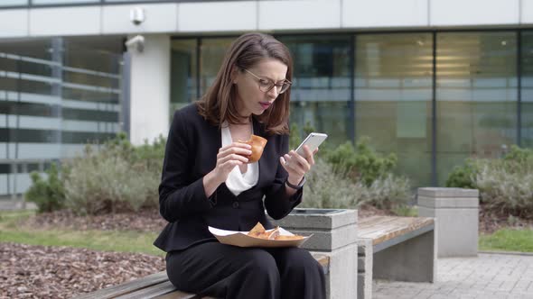 Businesswoman Browsing Smartphone and Snacking with Sandwich in Yard
