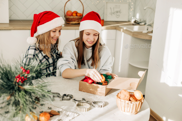 Sisters in Santa Claus hats are sitting at the kitchen table and packing sweet gifts to friends