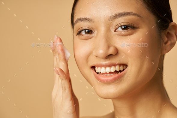 Studio portrait of lovely young woman smiling at camera while applying gentle foam facial cleanser