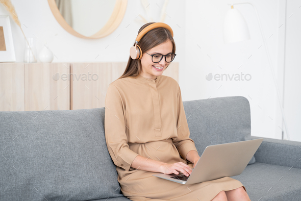 Young female listen to voice call through wireless headphones, sitting on couch with laptop