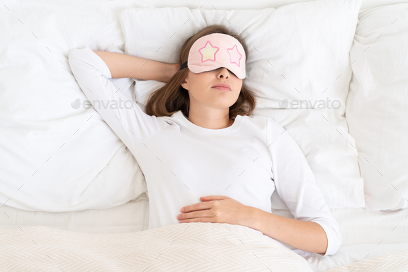 Young female dreaming in white bed under blanket in morning, wearing sleeping mask